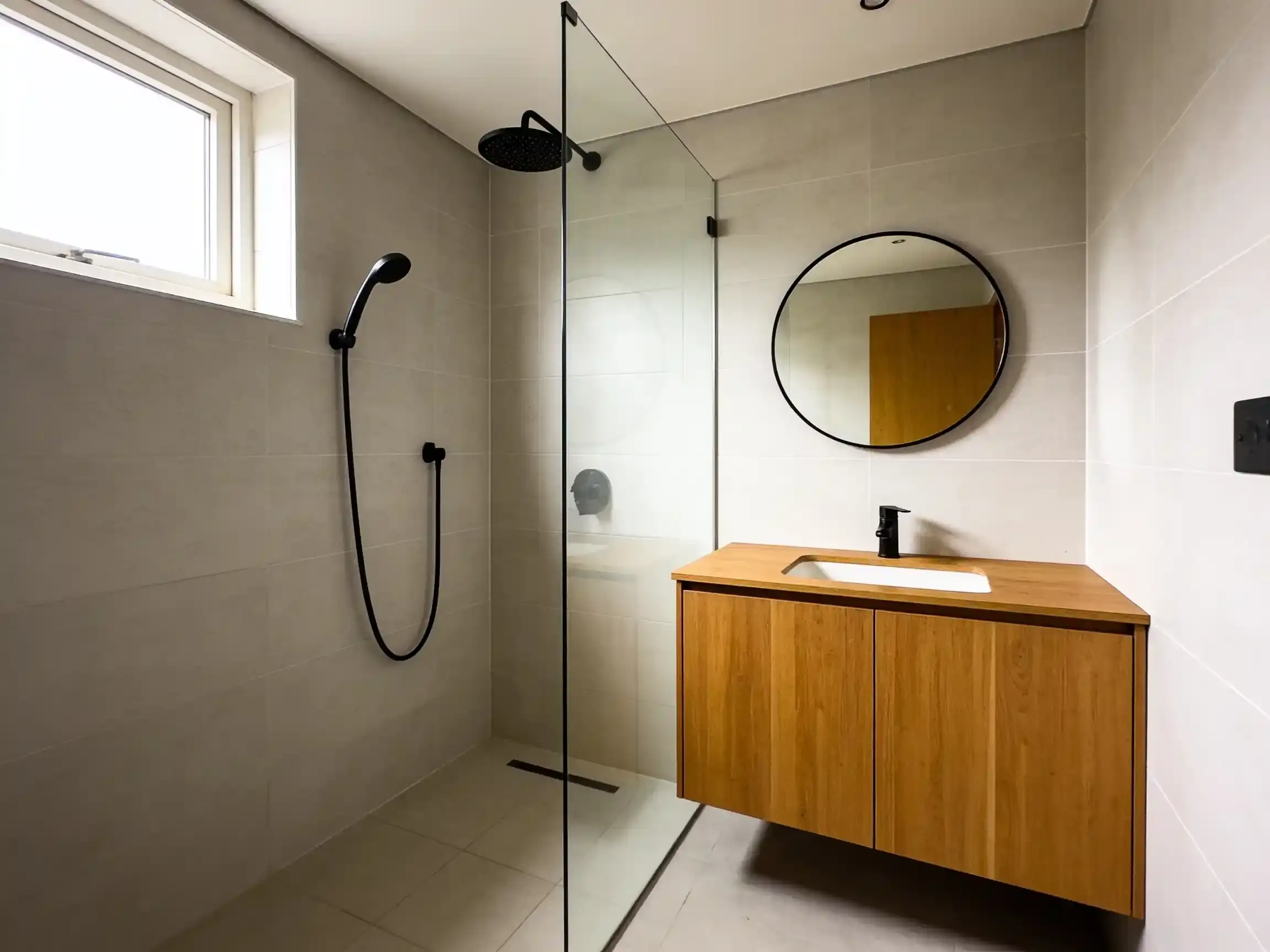 The same bathroom renovated with light grey tiles to the ceiling, a walk-in shower behind a frameless glass panel, a round mirror and a floating timber vanity