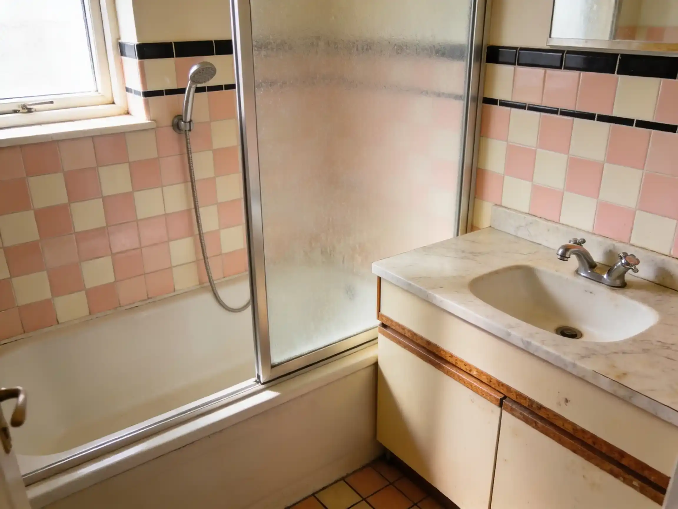A dated bathroom with pink and cream tiles, a shower over the bath behind a frosted framed screen, and a vanity with chrome twin taps