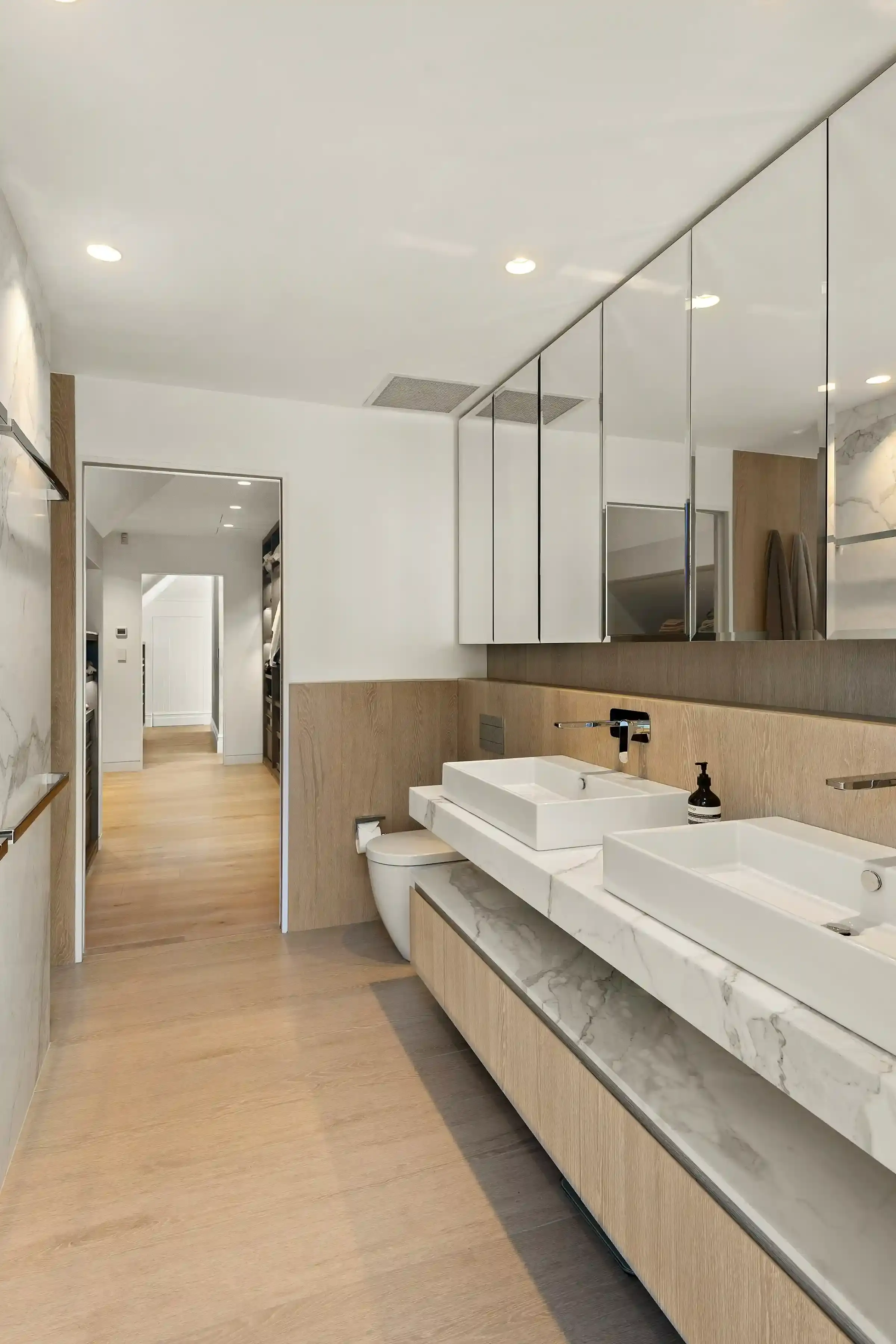 A long bathroom with timber-look floor tiles, a white double vanity with two vessel basins and a wall of mirrored cabinets