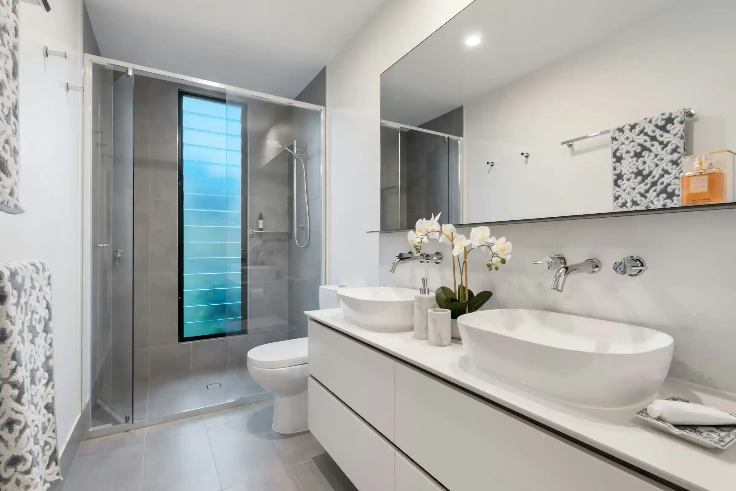 A bathroom with a white vanity and an oval vessel basin, a wall-mounted mixer, a frameless shower and a tall louvre window