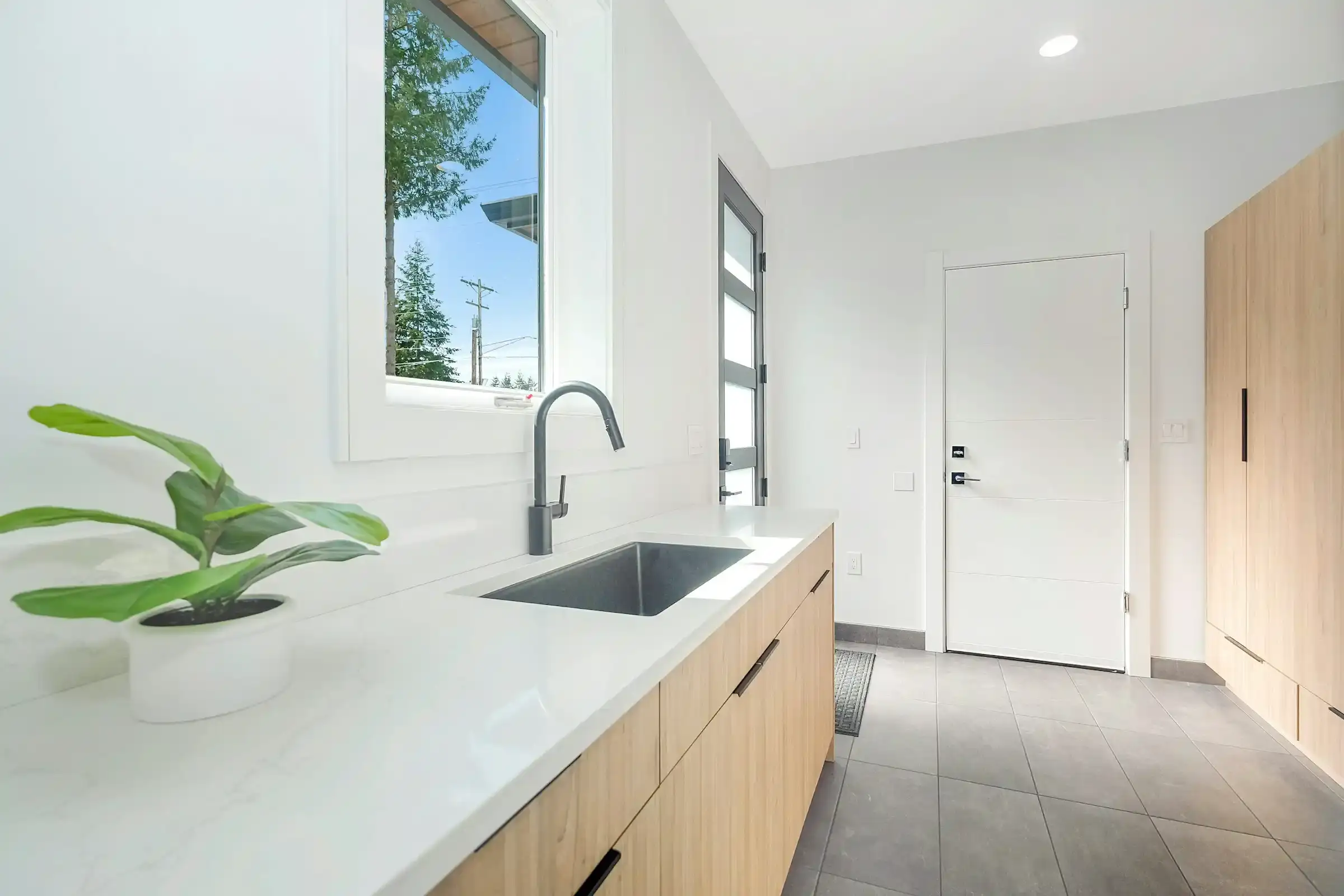 A laundry with a white stone benchtop, a stainless sink and a gooseneck mixer above timber-look cupboards, a plant on the bench