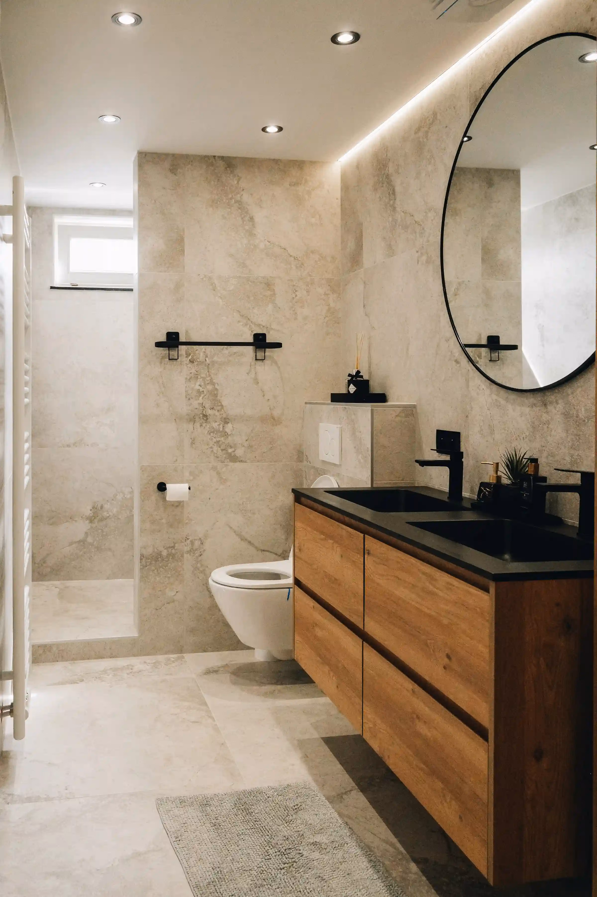 A renovated bathroom with stone-look tiles, a walk-in shower, a round mirror, a wall-hung toilet and a dark timber vanity with a black basin